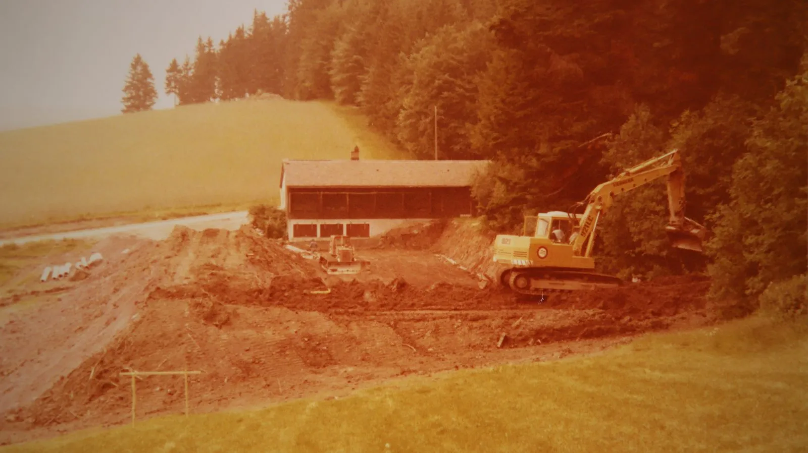 Farbaufnahme der Bauarbeiten: ein Bagger schiebt Erde zu einem Wall auf, dahinter das Schützenhaus mit rotem Dach, ringsum Wiese und Wald.