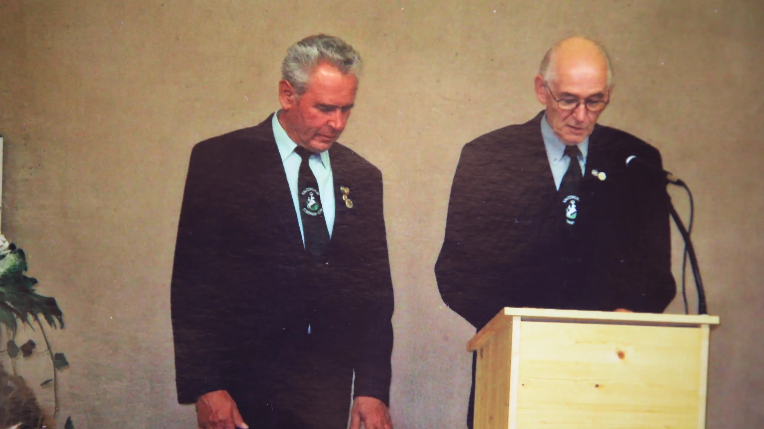 Farbaufnahme: zwei Männer in dunklen Anzügen mit Vereinsabzeichen stehen nebeneinander an einem hellen Holzpult.