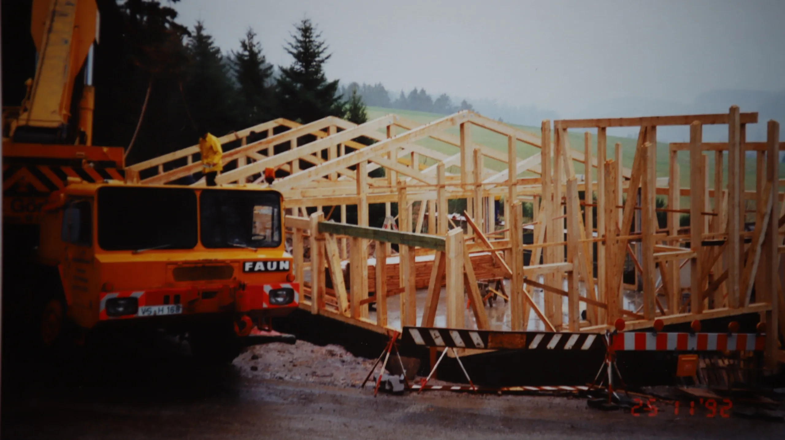 Farbaufnahme: ein Autokran hebt einen vorgefertigten Holzbinder über die entstehende Halle, im Vordergrund liegen weitere Binder bereit.
