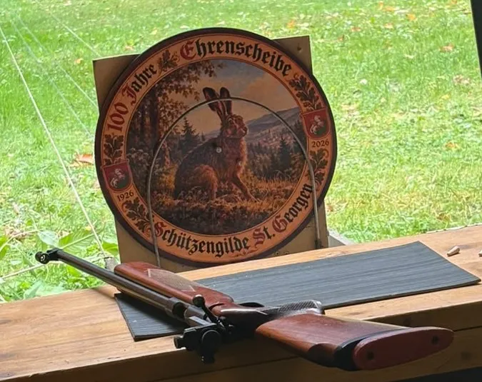 Die runde Ehrenscheibe zum Jubiläum mit einem Hasen in herbstlicher Landschaft und der Aufschrift „100 Jahre Ehrenscheibe, Schützengilde St. Georgen, 1926 2026", davor ein Luftgewehr auf dem Schießtisch.