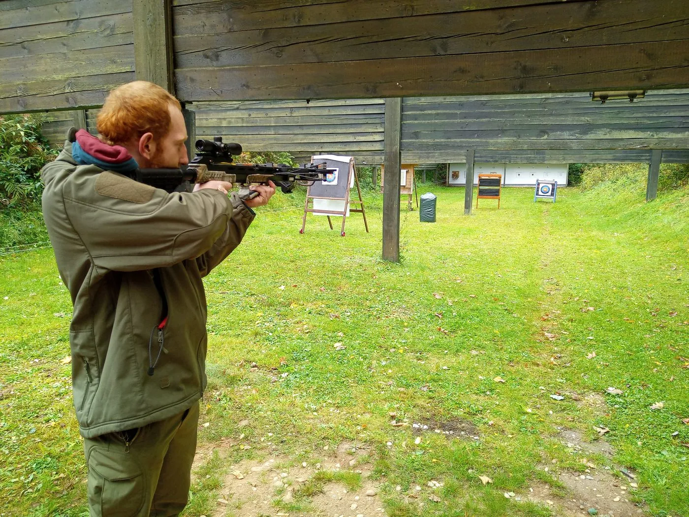 Ein Schütze steht am Außenstand und legt mit der Armbrust an; vor ihm auf der Wiese stehen Zielscheiben auf Ständern, dahinter die Holzverkleidung des Schützenhauses.