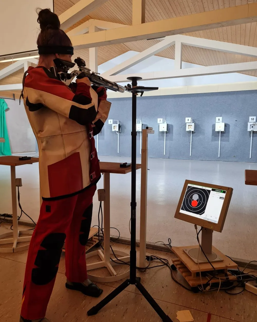 Eine Schützin im roten Schießanzug steht am 10-Meter-Stand und legt mit dem Luftgewehr an; daneben ihr Stativ mit der Trefferanzeige, im Hintergrund die Reihe der Scheiben unter dem Holzdach der Halle.