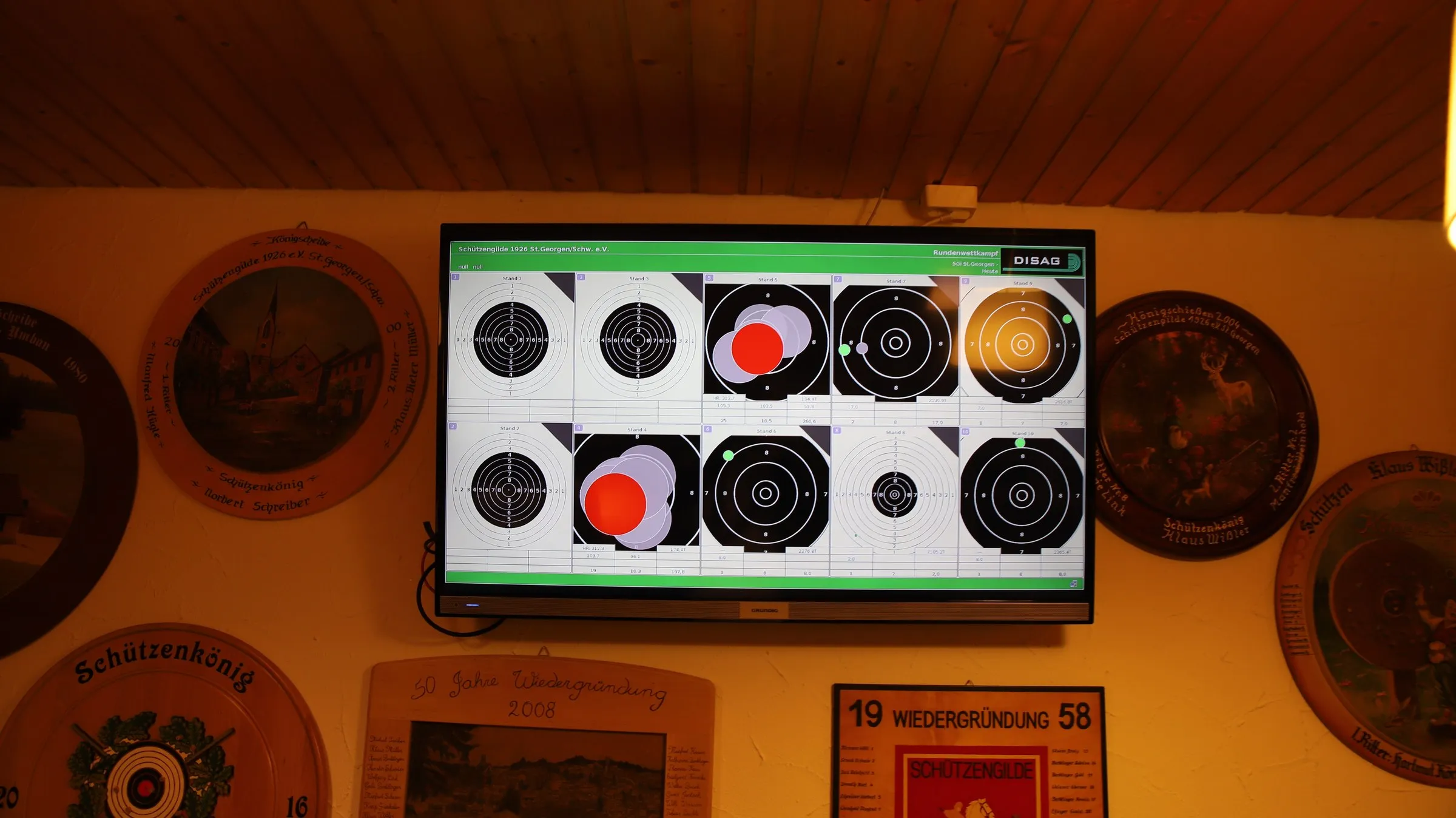 Ein Bildschirm an der Wand der Gaststätte zeigt zehn Schießscheiben mit den Treffern eines laufenden Wettkampfs, ringsum hängen bemalte Schützenscheiben.