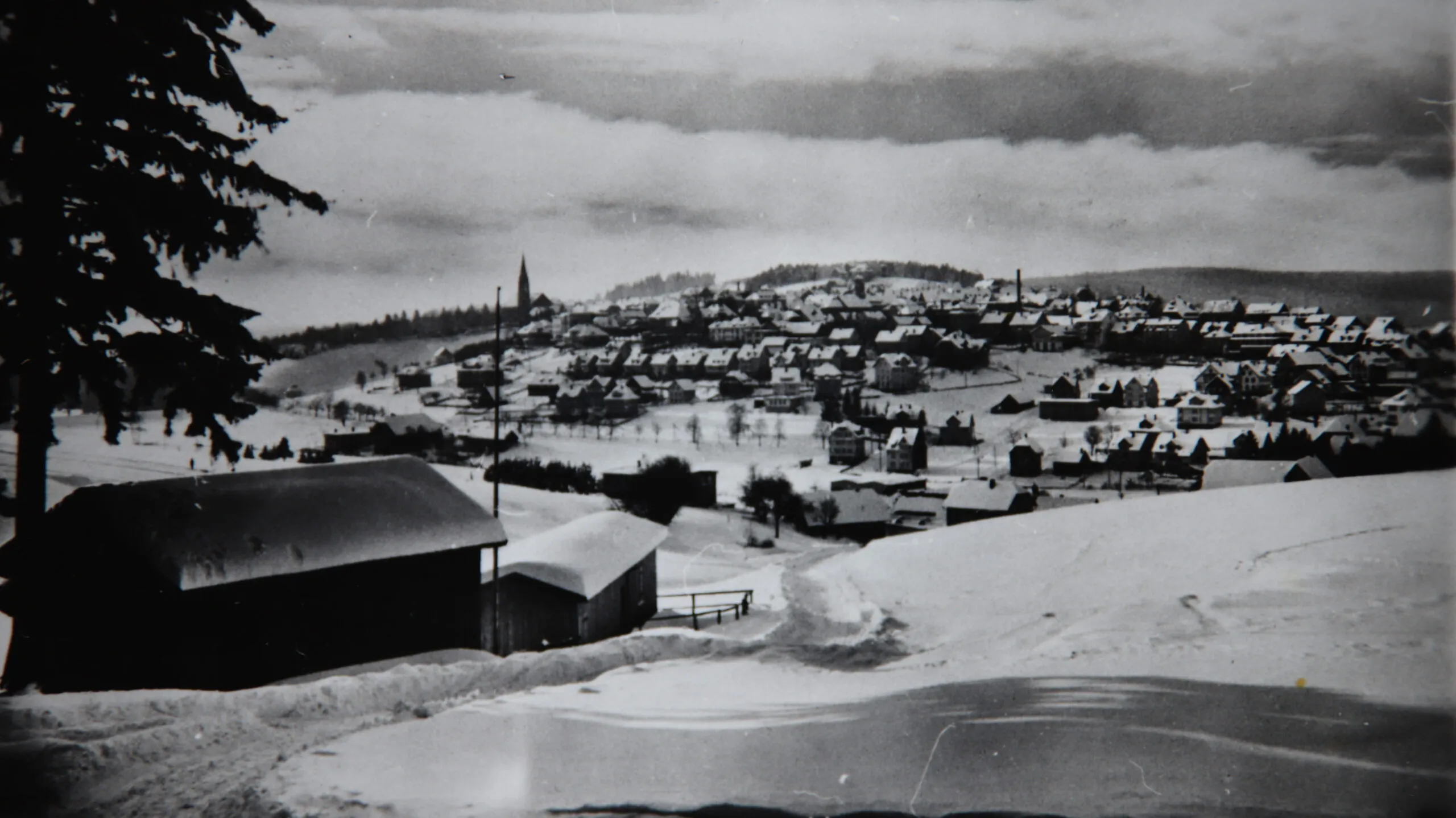Schwarzweißaufnahme im Winter: zwei dunkle Holzgebäude am verschneiten Hang, das linke lang und mit Satteldach, das rechte kleiner; dahinter liegt St. Georgen unter Schnee.