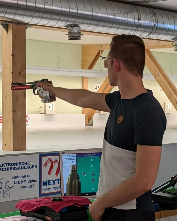 Ein Schütze steht am Pistolenstand und streckt den Arm mit der Luftpistole zur Scheibe aus; hinter ihm die Auflage mit Koffer und Zubehör.