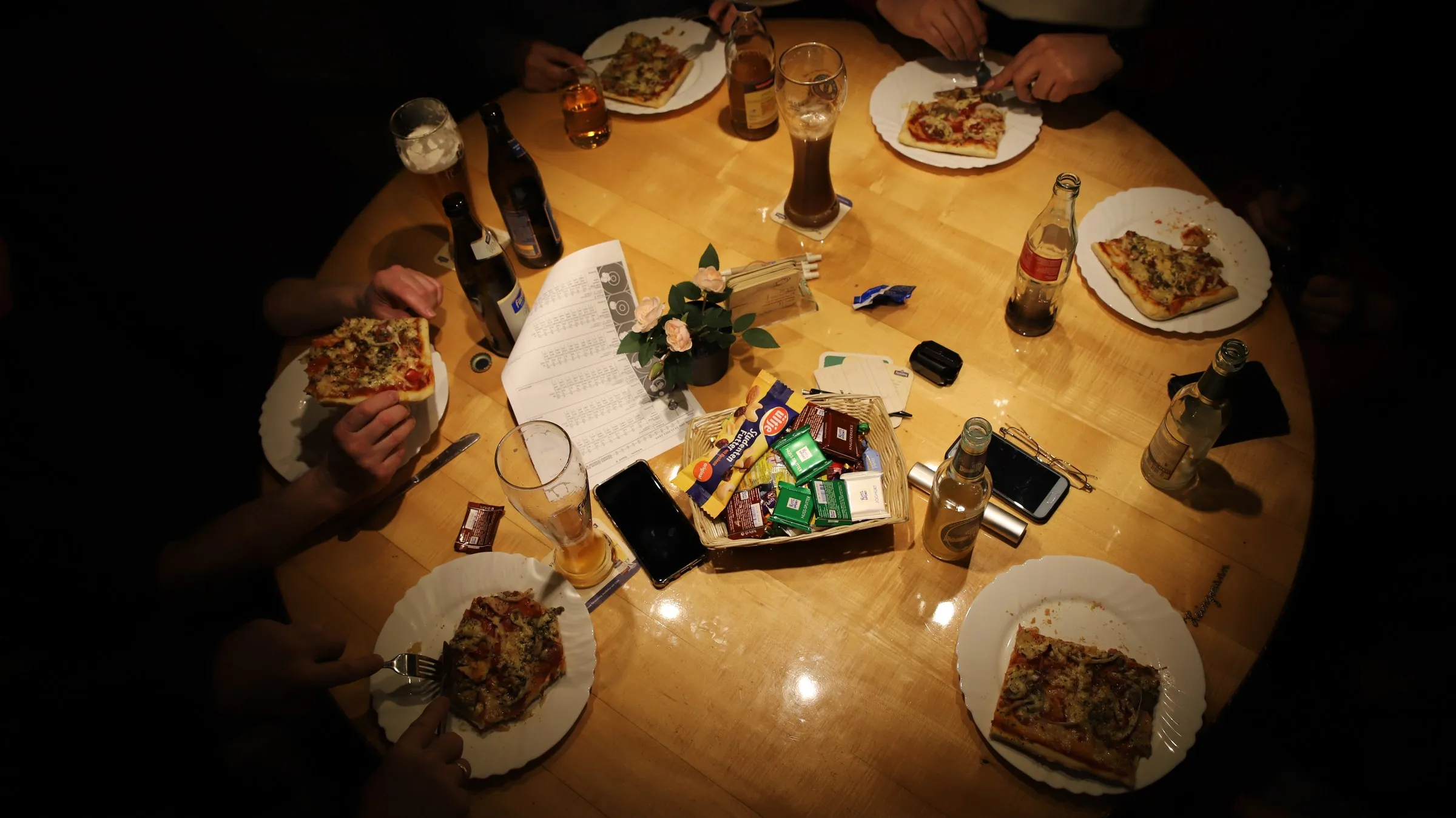 Blick von oben auf einen runden Stammtisch: Teller mit Pizza, Gläser und Flaschen, ringsum Hände von etwa sechs Personen.