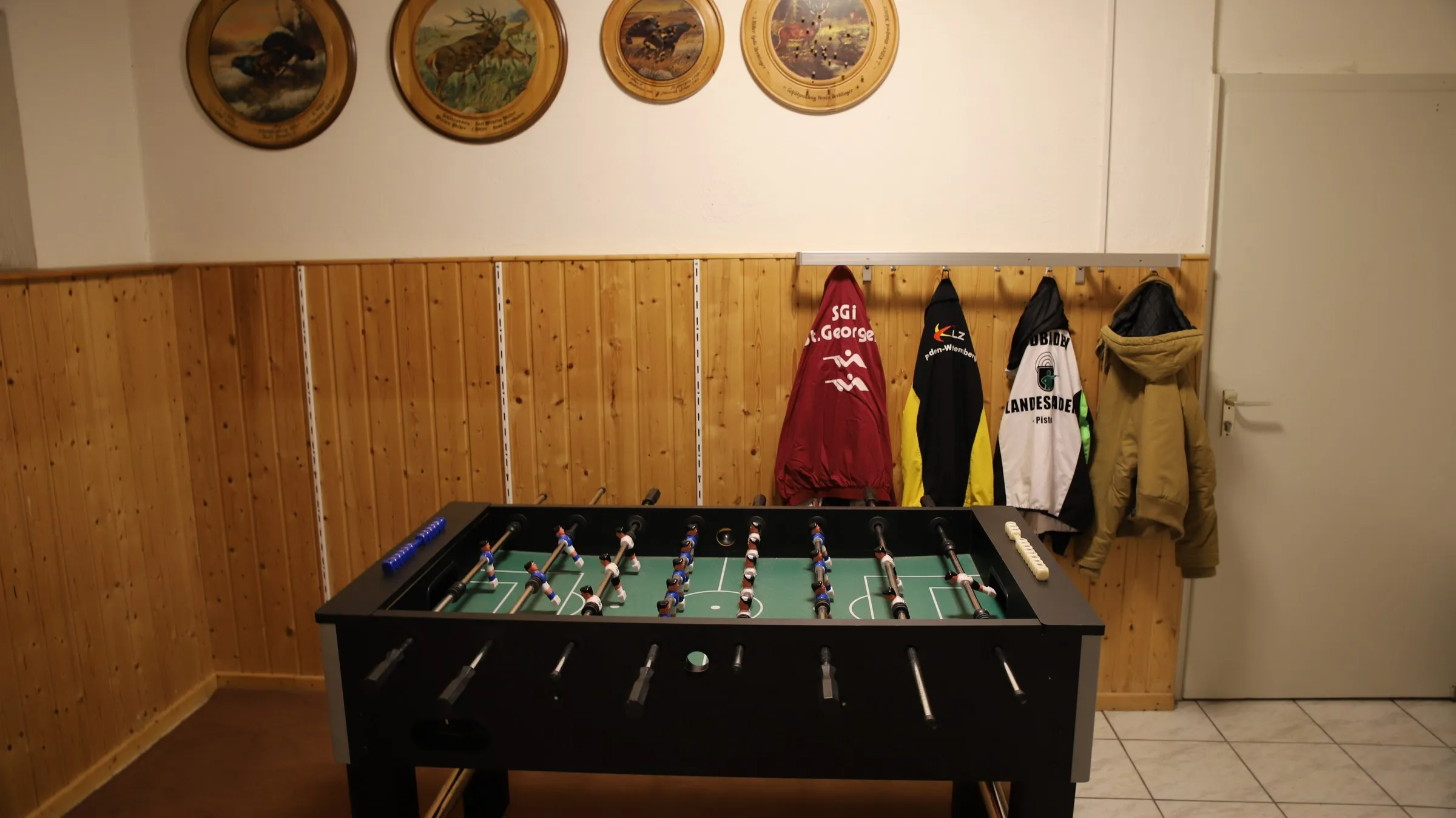 Ein Tischkicker steht in einem holzvertäfelten Raum, daneben hängen Vereinsjacken an einer Garderobe.